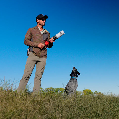 Man in outdoor clothing with a dog in a grassy field under a clear blue sky holding launcher
